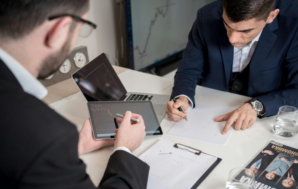 Two businessmen analyzing cryptocurrency market trends using a tablet and laptop in an office.